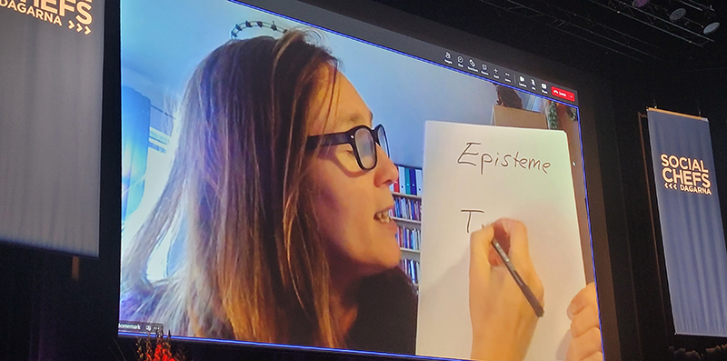 Kvinna med glasögon i Teamssändning håller upp papper framför kameran och skriver "Episteme".