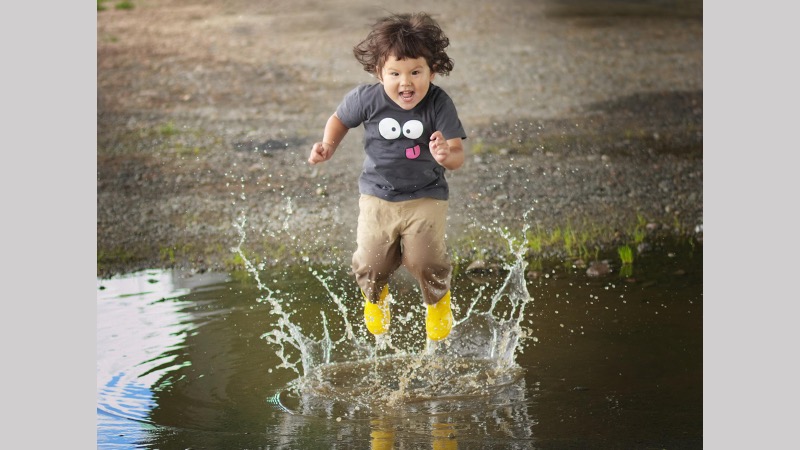 Ett skrattande barn med gula gummistövlar som hoppar högt i en vattenpöl